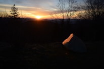 Sunset over a mountain range with a small group setting up camp, tents glowing warmly.