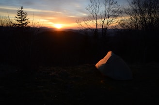 A serene sunset over a quiet forest campsite with a lone tent and a softly glowing campfire.