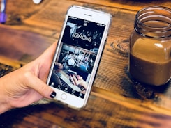 A hand holding a smartphone with a partially viewed app open, displaying sections labeled 'SERMONS', 'EVENTS', and 'GIVE'. The background is a wooden surface holding a small jar containing a brown beverage.