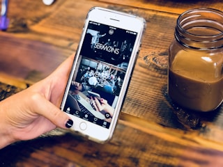 A hand holding a smartphone with a partially viewed app open, displaying sections labeled 'SERMONS', 'EVENTS', and 'GIVE'. The background is a wooden surface holding a small jar containing a brown beverage.