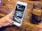 A hand holding a smartphone with a partially viewed app open, displaying sections labeled 'SERMONS', 'EVENTS', and 'GIVE'. The background is a wooden surface holding a small jar containing a brown beverage.