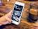 A hand holding a smartphone with a partially viewed app open, displaying sections labeled 'SERMONS', 'EVENTS', and 'GIVE'. The background is a wooden surface holding a small jar containing a brown beverage.