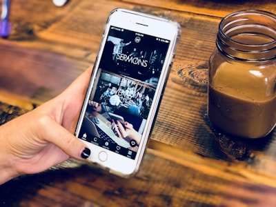 A hand holding a smartphone with a partially viewed app open, displaying sections labeled 'SERMONS', 'EVENTS', and 'GIVE'. The background is a wooden surface holding a small jar containing a brown beverage.