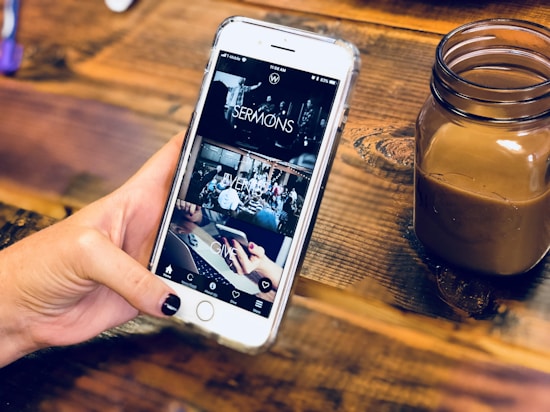 A hand holding a smartphone with a partially viewed app open, displaying sections labeled 'SERMONS', 'EVENTS', and 'GIVE'. The background is a wooden surface holding a small jar containing a brown beverage.