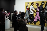 People are observing art in a gallery, with a large, colorful painting of stylized figures walking, set against a bright yellow background. Several individuals are standing and looking around, some engaged with phones or holding notepads. There is a modern art sculpture of interconnected globes to the side.