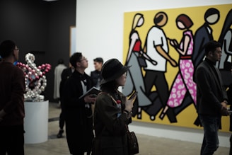 People are observing art in a gallery, with a large, colorful painting of stylized figures walking, set against a bright yellow background. Several individuals are standing and looking around, some engaged with phones or holding notepads. There is a modern art sculpture of interconnected globes to the side.