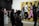 People are observing art in a gallery, with a large, colorful painting of stylized figures walking, set against a bright yellow background. Several individuals are standing and looking around, some engaged with phones or holding notepads. There is a modern art sculpture of interconnected globes to the side.