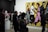 People are observing art in a gallery, with a large, colorful painting of stylized figures walking, set against a bright yellow background. Several individuals are standing and looking around, some engaged with phones or holding notepads. There is a modern art sculpture of interconnected globes to the side.