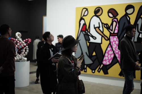 People are observing art in a gallery, with a large, colorful painting of stylized figures walking, set against a bright yellow background. Several individuals are standing and looking around, some engaged with phones or holding notepads. There is a modern art sculpture of interconnected globes to the side.
