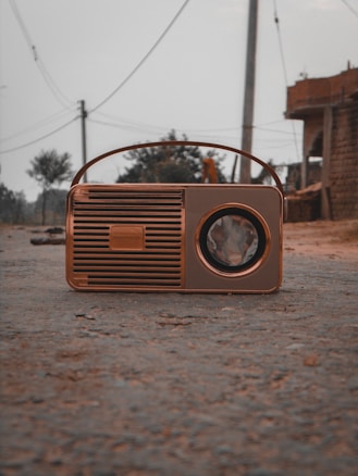 A vintage portable radio with a brown casing lies on a dirt road. In the background, there are utility poles and wires, with a partial view of a brick building and some trees.