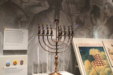 A menorah with small replicas of the Statue of Liberty's torch holders stands prominently. Informative displays are present around it, providing historical context. In the background, a detailed mural depicts figures engaged in various discussions, while a vintage poster promotes unity with an American flag illustration, emphasizing cultural convergence.