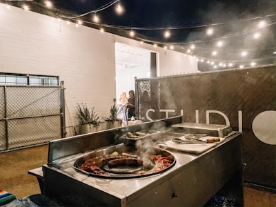 An outdoor setting with a taco stand featuring a grill filled with cooking sausages and other ingredients. String lights hang above, creating a warm and cozy atmosphere. A fence and potted plants are visible in the background, alongside a sign partially displaying the word 'STUDIO'. Two people are standing near the entrance, appearing engaged in conversation.