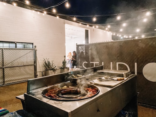 An outdoor setting with a taco stand featuring a grill filled with cooking sausages and other ingredients. String lights hang above, creating a warm and cozy atmosphere. A fence and potted plants are visible in the background, alongside a sign partially displaying the word 'STUDIO'. Two people are standing near the entrance, appearing engaged in conversation.