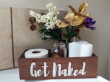 A wooden box with the phrase 'Get Naked' in white letters contains a roll of toilet paper, two white pillar candles, and a glass jar filled with small pinecones. It is decorated with an arrangement of artificial flowers, including white berries, gold leaves, purple and red blooms, and small brown pinecones. The background consists of beige tiled walls.