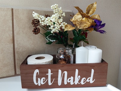 A wooden box with the phrase 'Get Naked' in white letters contains a roll of toilet paper, two white pillar candles, and a glass jar filled with small pinecones. It is decorated with an arrangement of artificial flowers, including white berries, gold leaves, purple and red blooms, and small brown pinecones. The background consists of beige tiled walls.
