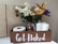 A wooden box with the phrase 'Get Naked' in white letters contains a roll of toilet paper, two white pillar candles, and a glass jar filled with small pinecones. It is decorated with an arrangement of artificial flowers, including white berries, gold leaves, purple and red blooms, and small brown pinecones. The background consists of beige tiled walls.