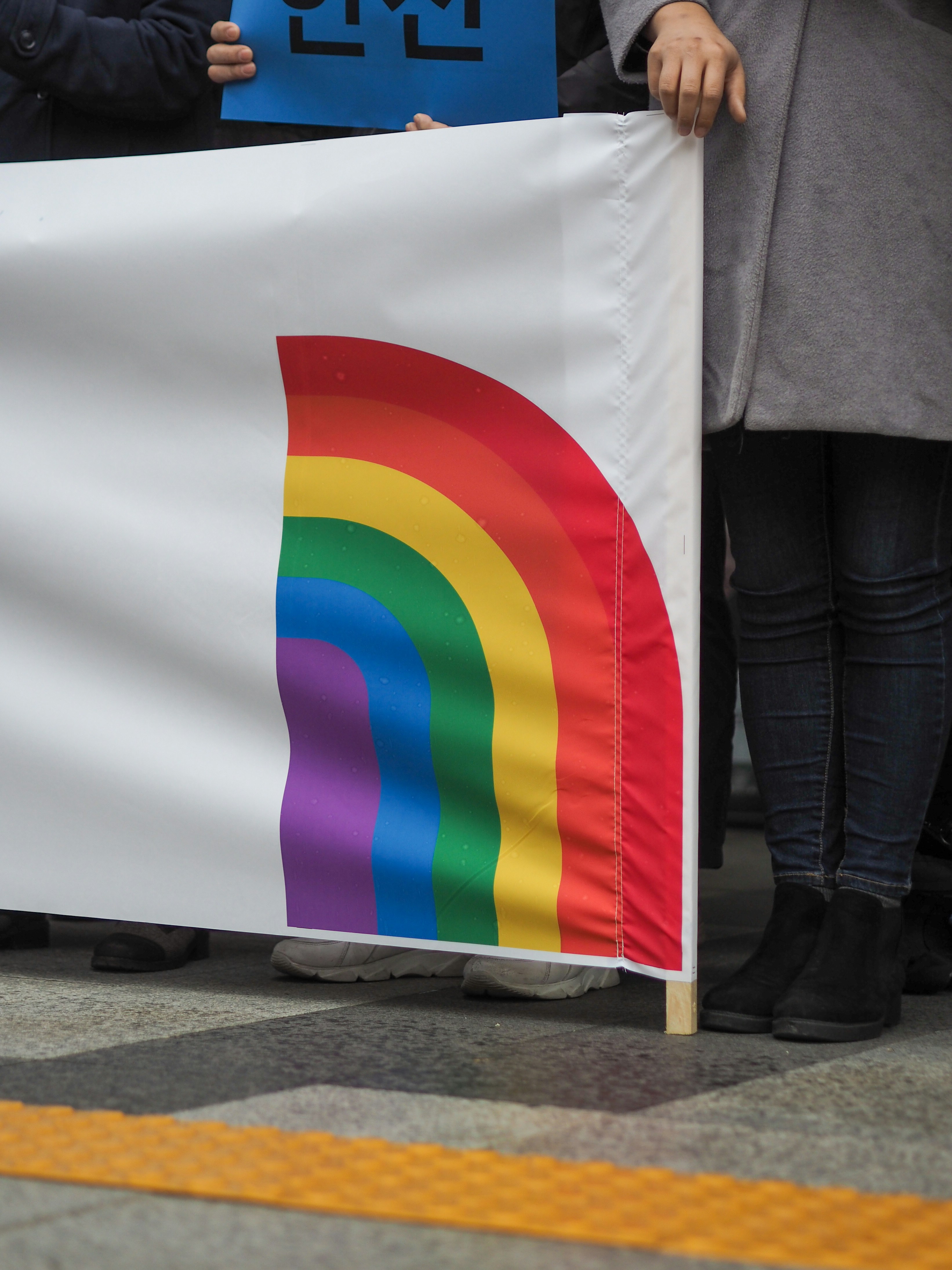 People holding a white banner with a rainbow design at a public gathering, representing LGBTQ+ visibility.