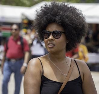 A person with an afro hairstyle and sunglasses is in a sunny outdoor setting. The individual is wearing a black top and a crossbody bag. In the background, there are blurred images of people walking and a white tent structure.