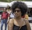 A person with an afro hairstyle and sunglasses is in a sunny outdoor setting. The individual is wearing a black top and a crossbody bag. In the background, there are blurred images of people walking and a white tent structure.