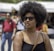 A person with an afro hairstyle and sunglasses is in a sunny outdoor setting. The individual is wearing a black top and a crossbody bag. In the background, there are blurred images of people walking and a white tent structure.