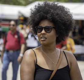 A person with an afro hairstyle and sunglasses is in a sunny outdoor setting. The individual is wearing a black top and a crossbody bag. In the background, there are blurred images of people walking and a white tent structure.