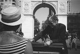 In a bustling urban setting, two individuals engage in a musical performance near a grand stone archway. One person, wearing a wide-brimmed straw hat and striped clothing, faces away while the other, dressed in a pinstripe suit and sunglasses, stands near a piano. This lively scene unfolds in a public square with tall buildings and people milling about in the background.