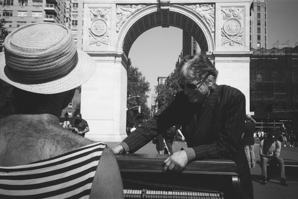 In a bustling urban setting, two individuals engage in a musical performance near a grand stone archway. One person, wearing a wide-brimmed straw hat and striped clothing, faces away while the other, dressed in a pinstripe suit and sunglasses, stands near a piano. This lively scene unfolds in a public square with tall buildings and people milling about in the background.