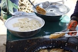 A person is cooking food outdoors, using a pair of scissors to handle a fried item. A large bowl filled with a batter or liquid containing slices of plantain or similar fruit is present. There's also a frying pan filled with oil, where the food is being cooked. A strainer with additional pieces sits nearby, and some utensils are on or beside the table.