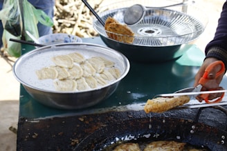 A person is cooking food outdoors, using a pair of scissors to handle a fried item. A large bowl filled with a batter or liquid containing slices of plantain or similar fruit is present. There's also a frying pan filled with oil, where the food is being cooked. A strainer with additional pieces sits nearby, and some utensils are on or beside the table.