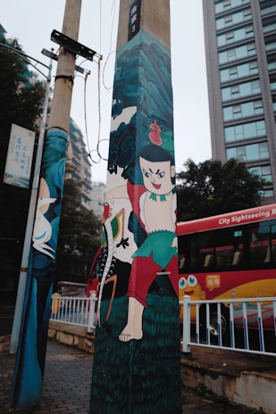 A street scene features a utility pole with vibrant cartoon-style artwork depicting characters with bold expressions and colorful clothing. In the background, a red city sightseeing bus is visible alongside modern buildings and trees.