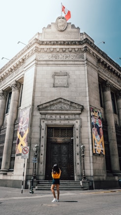 A grand, historical building with ornate stonework, flags raised at the top, and large banners on either side of the entrance reading 'Museo'. A person stands in front of the entrance, looking up at the facade.