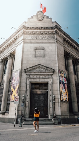 A grand, historical building with ornate stonework, flags raised at the top, and large banners on either side of the entrance reading 'Museo'. A person stands in front of the entrance, looking up at the facade.