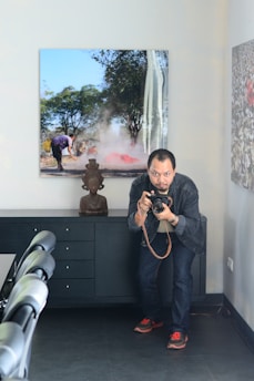 A person crouches indoors holding a camera, positioned in front of a dark wooden cabinet with drawers. On top of the cabinet sits a small sculpture of a head. The room contains an image on the wall depicting a person working in a smoky outdoor scene with trees in the background.
