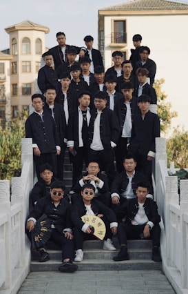 A large group of young men wearing similar traditional black outfits with white shirts are gathered on a set of stone steps. Some of them are holding fans with inscriptions. The setting is outdoors with a building visible in the background, and trees surrounding the area.