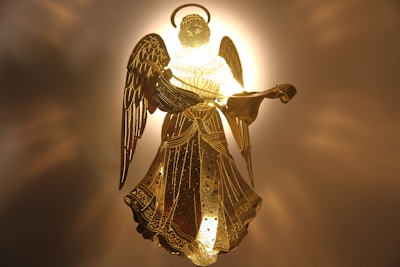 An intricately designed golden angel figurine is illuminated against a radiant backdrop, highlighting its detailed wings and flowing gown. The light creates a halo effect around its head, enhancing the celestial appearance.