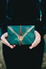 A pair of hands holds a decorative, green envelope secured with a leather strap and a wax seal, set against a dark background. The envelope features a subtle V-pattern and an elegant design.