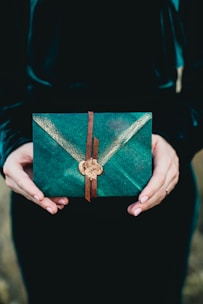 A pair of hands holds a decorative, green envelope secured with a leather strap and a wax seal, set against a dark background. The envelope features a subtle V-pattern and an elegant design.