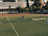 A football field with players scattered around, some gathered in a small group in the center, engaging in practice. The field is surrounded by trees and buildings in the background, with a clear sky above. Two individuals stand on the sidelines while others are positioned near soccer balls on the field.