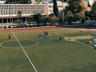 A football field with players scattered around, some gathered in a small group in the center, engaging in practice. The field is surrounded by trees and buildings in the background, with a clear sky above. Two individuals stand on the sidelines while others are positioned near soccer balls on the field.