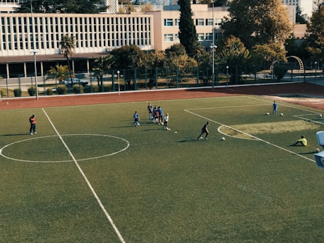 A football field with players scattered around, some gathered in a small group in the center, engaging in practice. The field is surrounded by trees and buildings in the background, with a clear sky above. Two individuals stand on the sidelines while others are positioned near soccer balls on the field.