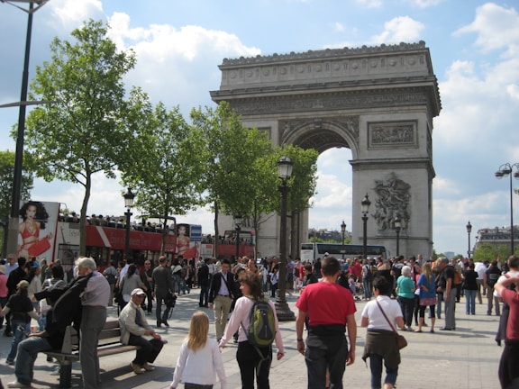 A large, iconic arch stands prominently in a bustling public area filled with people walking and gathering. Bright green trees line the area alongside advertisements visible on a red tour bus. The sky is partially cloudy, casting soft shadows on the scene below.