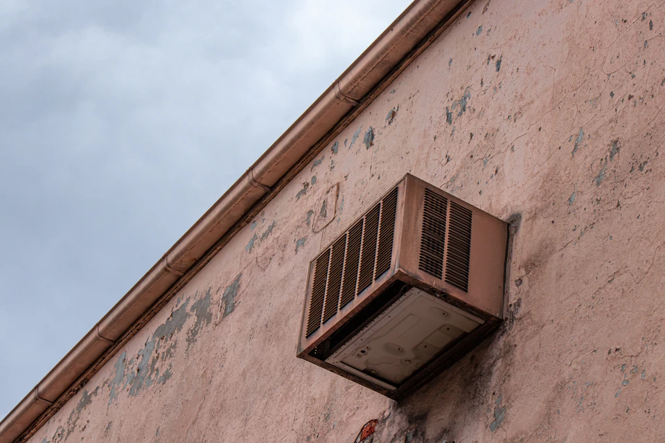 The image shows a brown building with a large window on its side, which is currently closed and attached to the wall by brackets. The window appears to be an air conditioning unit or heat pump, depending on the season. The building has a faded pinkish-brown exterior that suggests it may be old or exposed to the elements for some time.

The sky in the background is filled with clouds, indicating an overcast day. The image does not contain any discernible text or human figures, and there are no other objects present besides the window on the building.