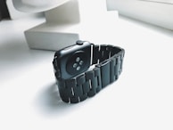A black smartwatch with a metal link band is placed on a white surface near a window, allowing natural light to emphasize its sleek design. The smartwatch has a dark screen and is angled to show the sensors on its underside.