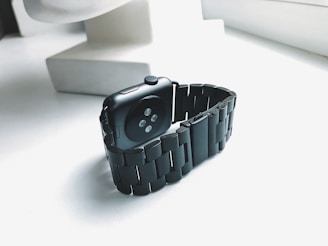 A black smartwatch with a metal link band is placed on a white surface near a window, allowing natural light to emphasize its sleek design. The smartwatch has a dark screen and is angled to show the sensors on its underside.
