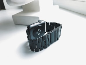 A black smartwatch with a metal link band is placed on a white surface near a window, allowing natural light to emphasize its sleek design. The smartwatch has a dark screen and is angled to show the sensors on its underside.
