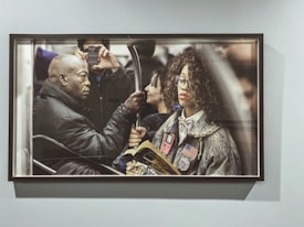 A framed photograph depicts a crowded subway scene. A woman with curly hair, wearing glasses and a coat adorned with patches including an American flag, is holding a book. Beside her, a man in a black jacket is capturing a photo with his smartphone. The image conveys the hustle and bustle of urban commuting.