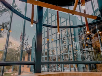 A modern interior space with large glass windows and visible metal beams. Wooden light fixtures are suspended from the ceiling, and there are decorative line drawings of birds on the glass. Outside, a building with brick walls and trees can be seen.