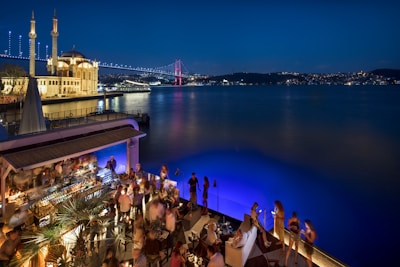 A lively rooftop bar overlooking a wide river at night. The scene is bustling with people socializing near a well-lit bar area. In the background, an illuminated mosque stands prominently, and a bridge adorned with lights spans across the river. City lights shimmer in the distance, adding to the vibrant ambiance.