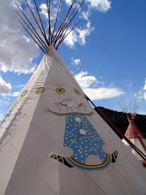 A large tipi with traditional Native American designs stands under a vibrant blue sky. The tipi features artistic depictions with colorful geometric patterns and symbolic imagery. In the background, another tipi is visible, along with fluffy white clouds.
