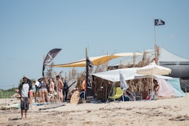 A beach scene with a group of people under makeshift tents and umbrellas. Surfboards and flags are visible, with a pirate flag prominently displayed. The background shows tall grasses and the sky is clear.